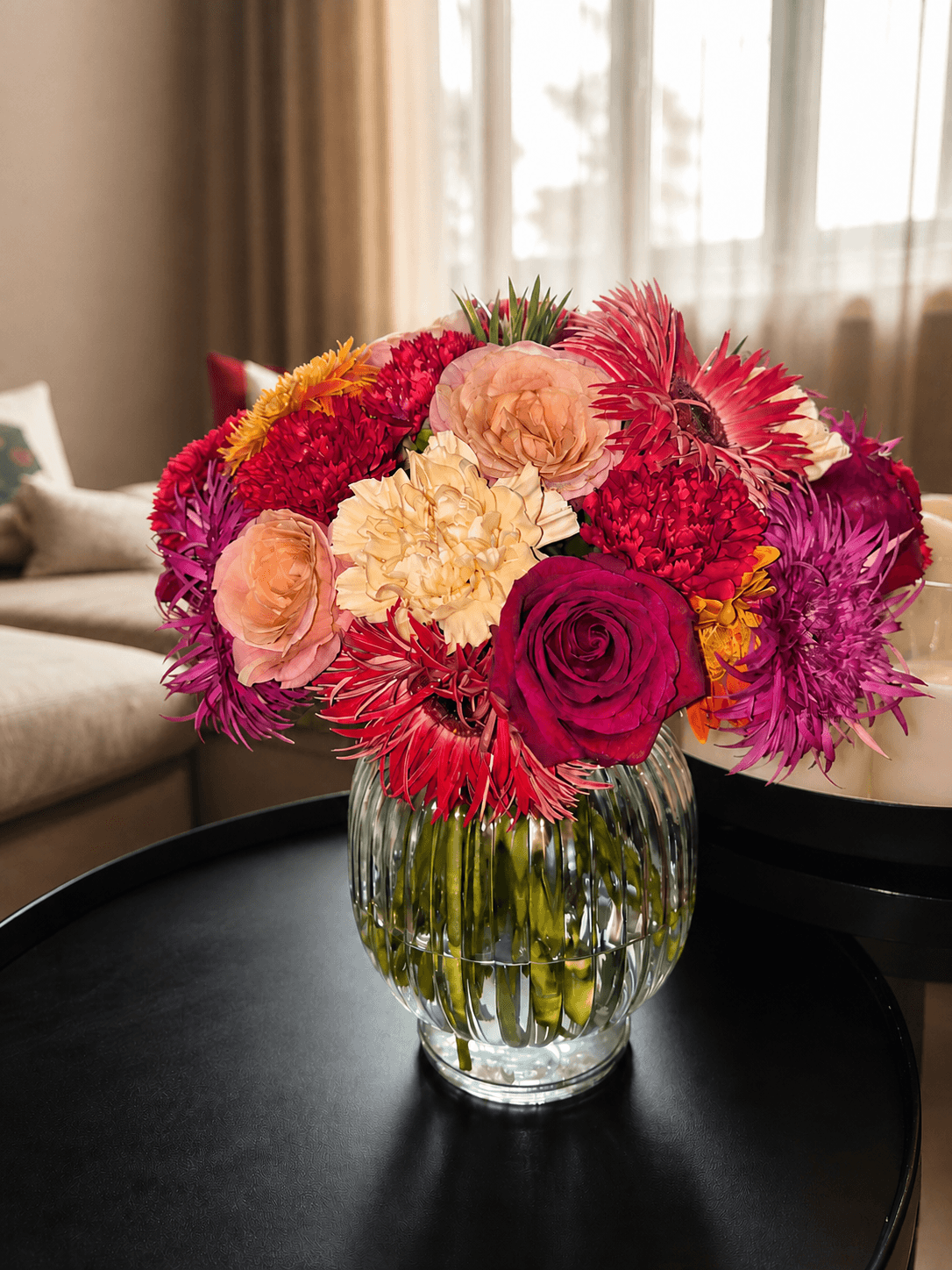 Vibrant pink, red, coral and cream floral arrangement in a clear ribbed vase on a black table.