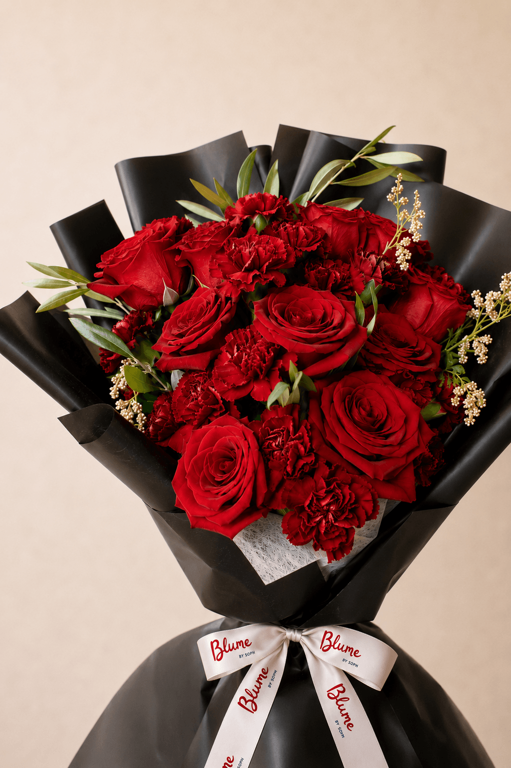 Deep red roses and carnations with green foliage, wrapped in black paper and finished with a white Blume ribbon.